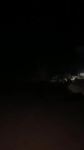 The media file appears to be a collection of short video clips taken at night. The initial clips show a dimly lit outdoor scene with a gathering of people. In the background, there are lights from buildings and possibly a tower with a red light. The subsequent clips focus on a person in the foreground, with their hand and head visible. The background in these clips remains dark, with some distant lights visible. The overall mood seems somewhat subdued due to the darkness and limited visibility. The location is identified as Shybartubek, Kazakhstan, and the time is clearly nighttime. There are no visible text elements within the media.