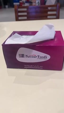 The video shows a tissue box on a table. The tissue box is pink and purple and has the logo for "Savour Foods" on it. There are tissues coming out of the top of the box. In the background, there is a wooden bench with people sitting on it. The table appears to be in a restaurant or food court. The video is static and there are no visible actions or interactions.