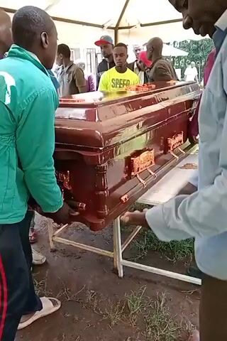 Here is a concise description of the video:

The video depicts a funeral scene. A shiny, dark-wood casket rests on a metal stand beneath a white tent. Several people stand around the casket. Some are dressed casually, others in more formal attire. There is a camera and tripod visible in the background. The setting appears to be outdoors, on a grassy area. The atmosphere is somber.