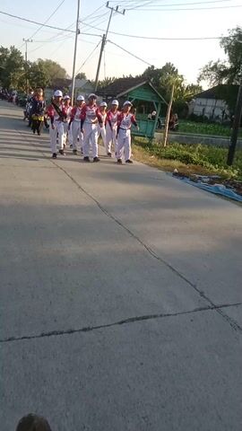 A group of people in uniform are lined up on a road. They are wearing white pants, white shirts with red accents, and white hats. Some of them are holding their hands up in a salute. There are motorcycles parked on the side of the road, and some people are riding on them. The scene appears to be in a rural area, with houses and trees in the background. The sun is shining, and there are shadows on the road.