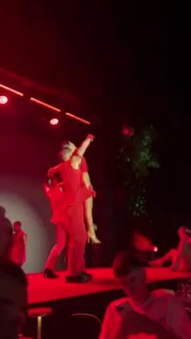 Here's a detailed description of the video:

**Setting:**
The video appears to be taken at a club or entertainment venue, possibly outdoors given the background of foliage on the right side of the screen. The lighting is predominantly red, creating a dramatic atmosphere. The camera is positioned from the audience's perspective, looking up at a stage.

**People:**
*   There is a group of performers, all dressed in red outfits. The men wear red vests and pants, with bare chests and biceps. The women wear red dresses.
*   There is at least one individual in the audience, visible in the foreground, wearing a white T-shirt and looking at their phone.

**Actions and Interactions:**
*   The performers are engaged in a dance routine.
*   Initially, one male dancer is holding a female dancer aloft.
*   The male dancers squat down and lift the female dancer above their heads, forming a sort of human pyramid.
*   Other female dancers are also performing movements on the stage.

**Objects:**
*   The stage is equipped with spotlights, creating a focused illumination on the performers.
*   There is a disco ball hanging behind the stage.
*   A neon sign is visible, displaying the letters "LU" in script.

**Overall Impression:**
The video captures a live performance, likely a dance or cabaret act, with a strong visual emphasis on the color red. The act involves coordinated movements, including acrobatic lifts and poses. The atmosphere seems energetic and vibrant.