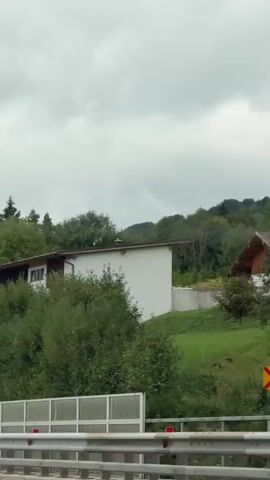 The video shows a view from inside a moving vehicle, likely on a highway, with a cloudy sky overhead. The road has a white barrier on the right side. On the left, there's a sign for Mondsee, indicating it's 700 meters away, and showing routes U62 and 264. Further down the road, a white truck with a red trailer is visible. The landscape includes trees and a small building on a hill. Another vehicle with bicycles on its rear is ahead of the truck. The scene appears to be in a rural or suburban area with lush green vegetation. There are no people clearly visible.