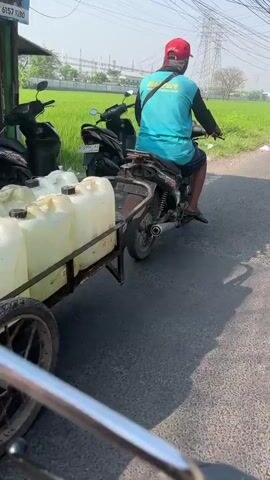 A person wearing a red cap, blue shirt, and dark shorts rides a motorcycle. The motorcycle is pulling a cart with multiple large, white jerrycans. The person is on a paved road with a green grassy field on the right side. In the background, there are power lines and a structure that appears to be a transmission tower. Several other motorcycles are parked to the left of the road. The video is shot from a slightly elevated, moving perspective, suggesting the videographer is also on a motorcycle. The lighting indicates it is daytime, likely sunny.