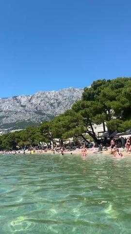 This is a short video captured at a beach on a sunny day. The water is clear and turquoise, with gentle ripples. In the foreground, the camera is low, appearing to be submerged slightly in the water, looking out towards the beach.

The beach is crowded with people sunbathing and swimming. Behind the beach, there is a line of lush green pine trees, and further behind them, a range of majestic, rocky mountains under a clear blue sky. Buildings are visible nestled amongst the trees and on the slopes of the mountains.

The video then cuts to a person in the water, with a white towel draped over their shoulders, looking off-camera. This is followed by a shot of a large, inflatable water park with slides and platforms, where people are visible playing and climbing. The text "peakz" is visible on the inflatable structure.

The overall impression is a vibrant, sunny beach vacation scene, likely in the late morning or early afternoon given the bright sunlight and shadows. The mood appears relaxed and joyful. The location is identified as Makarska, Croatia.