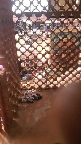 The video shows a view from what appears to be a building, looking out at a dusty, red dirt street scene. A wall made of interlocking circular clay objects partially obscures the view, but the street is visible through the holes. There are motorcycles parked and moving on the street, as well as people walking around. A large blue sign reads "DANGOTE CEMENT THIS WAY." The overall environment appears to be a small town or village, possibly in a rural area, with simple buildings visible in the background.
