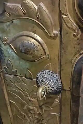 The video shows a close-up of a person's hand interacting with an ornate, golden door handle. The door appears to be made of metal with intricate carvings, including a large eye-like motif and swirling patterns. The handle itself is also decorative, with a round, textured base and a cylindrical grip. The hand, with visible fingernails and a red bracelet on the wrist, reaches for and turns the handle. The lighting suggests an indoor environment, possibly a temple or a place of historical significance due to the elaborate design of the door. The movement of the hand and handle is the primary action, indicating an attempt to open or operate the door.