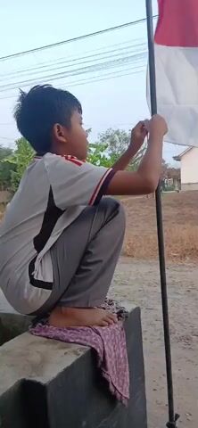 A young boy wearing a gray shirt with red and black accents and gray pants is squatting on a concrete ledge, with his right foot bare. He is tying a red and white flag to a black pole. The flag is partially unfurled and flapping in the wind. Behind the boy, there is dry grass, some trees, and a house. The sky is overcast.