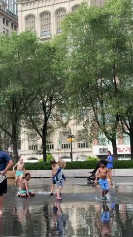 The media file captures a group of children playing in a public fountain on a bright, overcast day, likely in the early afternoon. The scene is set in an urban environment, with a large, ornate building, possibly a university or historical landmark, serving as the backdrop. Tall, lush trees flank the fountain, casting dappled shadows and suggesting a warm season, likely summer.

The main subjects are several children, mostly boys aged around 5-10 years old, all wearing swimwear. They are actively engaged in splashing water, running through the spray, and generally enjoying themselves in the shallow water. One child is seen sitting on the wet ground, while others are mid-stride, creating splashes.

Notable details include the wet, reflective surface of the fountain, mirroring the surrounding trees and the building. The weather appears to be mild, with no direct sunlight, and the sky is likely cloudy, contributing to the diffused lighting. The presence of urban architecture and manicured trees points to a well-maintained public space.

There is some visible text on signage in the background, including what appears to be part of a store name, "MACY'S," and potentially other retail or business establishments. The overall mood conveyed is one of joyous, carefree childhood play in an urban oasis. The children's expressions, though not clearly visible, can be inferred to be happy and energetic given their activity. The setting of Chicago is a plausible guess given the architectural style and the presence of such public water features designed for recreation.
