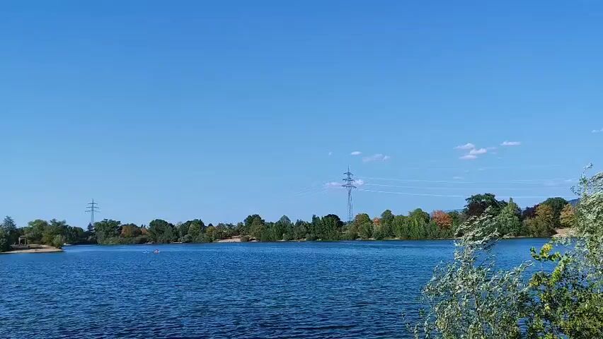 A wide shot captures a serene blue lake under a clear blue sky. The opposite shore is lined with a dense band of green and autumn-colored trees. In the background, several utility poles with power lines are visible, suggesting the presence of infrastructure near the natural landscape. The water appears calm with gentle ripples, reflecting the bright sky. On the lake, two small figures can be seen in what appear to be kayaks or canoes, suggesting recreational activity. The foreground features lush green foliage, with leaves rustling gently, adding a touch of movement to the otherwise still scene.