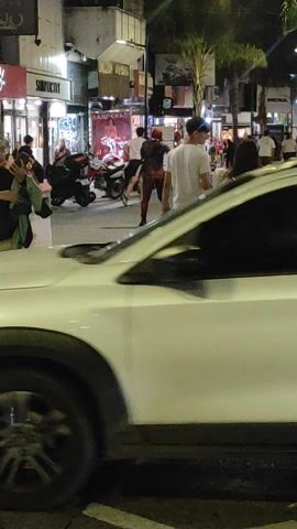 The video depicts a nighttime street scene in Lanús Este, Argentina. A white vehicle, possibly a car, is initially visible on the right, showcasing its rear red taillight and wheel. The camera then pans or moves to reveal a pedestrian area with several people. Two individuals are seen standing close together, possibly embracing. Further along the street, a person dressed in a red and black costume (resembling Deadpool) is captured in a dynamic pose, suggesting a performance or interaction. A motorcycle is parked nearby. The setting is an urban environment, characterized by illuminated storefronts and streetlights, indicating the late hour. The ground features a distinct patterned pavement with diagonal lines. Visible text on building signs includes "BANDERA S", "SIMPLICITY", and "RENA". The overall atmosphere is bustling and lively.