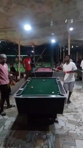 The video shows an outdoor pool hall with multiple pool tables and people playing or watching. The room is covered by a white, tent-like roof and is lit with multiple bright ceiling lights. The floor is a patchwork of different colored tiles.  Several men are visible in the video, some with pool cues in hand, moving around the tables. There are green pool tables in the foreground and a red pool table further back. Some individuals are sitting on the side. The video pans across the scene, showing the activity in the pool hall.