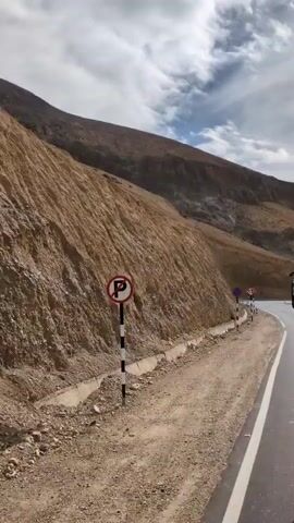 The video shows a roadside view from a moving vehicle on a mountain pass. The road is paved and has a white line marking its edge. On the left side of the road is a steep, barren mountainside with loose gravel and rocks. A concrete drainage ditch runs along the base of the mountain. Several road signs are visible. The first sign is a blue circle with a red X, indicating no stopping. Next is a sign with a black arrow curved to the left and then right, warning of a hairpin bend. Following that is a triangular sign with a wavy line, indicating a rough road. Another sign shows "20" in a red circle, indicating a speed limit of 20. Finally, there is a triangular sign with a black U-shaped arrow pointing down, indicating a U-turn. The sky is cloudy with patches of blue. The video is shot from a moving perspective, suggesting the vehicle is driving along the mountain road.