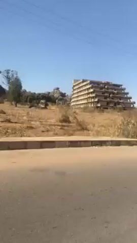 The video shows a building with a unique terraced design, positioned on a small hill with dry grass. The building is unfinished. The camera moves along a road, showing telephone poles, sparse trees, and the building in the distance. The sky is clear and blue. No people are visible.