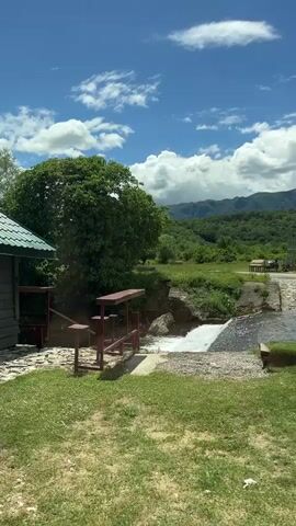 The video shows a grassy landscape with a small dam or waterfall. The water flows over the structure and into a pool below. A small building with a green roof is visible on the left. There is a wooden structure that appears to be for supporting something over the water. In the background, there are rolling hills covered in trees under a blue sky with puffy white clouds.