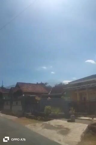 The video shows a street view of a residential area under a clear blue sky with sparse clouds. The camera is moving, indicating it might be from a vehicle. On the right side of the street, there are houses with red tile roofs and green walls, bordered by fences. A large tree with dense foliage is prominently visible on the right, with sunlight filtering through its leaves, causing some glare. On the left side of the street, there are more houses and fences. The road surface is asphalt, and there are some patches of dry grass and dirt along the roadside. The overall impression is of a sunny day in a suburban or rural neighborhood.