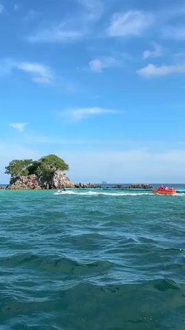 A small, rocky island with green trees grows out of the turquoise ocean under a bright blue sky with scattered white clouds. A red boat with several people on board speeds across the water to the right of the island. Several other people are visible on and around the island.