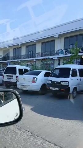 The video shows a parking lot in front of a commercial building with multiple businesses. Several cars are parked, including a white van and a white sedan. A white truck with a detached cab is being worked on by two men. Tents are set up in front of the building. The sky is clear and blue.