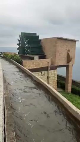 A stone canal carries a stream of water toward a green water wheel, which is housed within a light-brown stone structure. The water wheel slowly turns. The canal runs between stone walls, along which are some plants. The background features an overcast sky and a body of water with the horizon line visible. The structure and canal are elevated on what appears to be a cliff.
