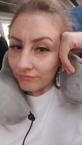 The video features a woman with light-colored hair, styled in a casual messy bun, looking directly at the camera in a selfie-style recording. She has a fair complexion, with defined eyebrows and visible long eyelashes. She wears a simple white crew-neck t-shirt and prominent silver-toned, multi-strand beaded earrings featuring white or clear beads. A gray U-shaped travel neck pillow is positioned around her neck, and a black cord, possibly from headphones or the pillow's drawstring, hangs down in front of her.

The setting appears to be inside a vehicle, likely a bus or train, indicated by the overhead paneling and a blue seat/headrest visible in the background. The lighting is internal and consistent.

Throughout the short clip, the woman displays subtle shifts in expression. She initially rests her head gently on her hand, looking towards the camera with a soft, slightly pensive smile. Around the 0:01 mark, her expression becomes more neutral or slightly serious, before returning to a faint, relaxed smile by 0:02. Her gaze is generally direct and engaging, conveying a sense of calm and self-awareness, perhaps a mix of contentment and slight tiredness from travel. The overall mood is quiet and introspective, consistent with being a passenger on a journey in Donji Jasenjani, Bosnia and Herzegovina.