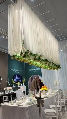 The camera pans across a display at a trade show. The initial focus is on a dining table set for a formal event, adorned with fine china, silverware, and floral arrangements. Above the table, a canopy of white fabric with floral garlands hangs suspended. A couple stands at the table. To the right, a booth for SONJA QUANDT SILBERMANUFAKTUR features silver objects and several people, some standing and some seated at high tables. The environment is spacious and well-lit with overhead track lighting.