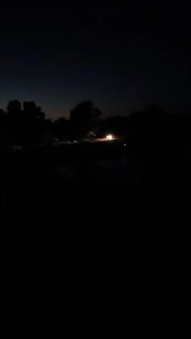 A car with its headlights on moves through a dark, rural landscape at dusk. The car is silhouetted against the faint glow of the horizon. Trees and foliage line the road, and a railroad track is visible in the foreground. A signal light with an orange glow can be seen in the distance.