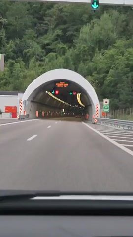 The video shows a car driving through a tunnel in Praveshka Lakavitsa, Bulgaria. The tunnel is well-lit with rows of lights along the ceiling and walls, and there are also red and green indicator lights. The road is paved and appears to be a highway, with lane markings visible.

A digital sign at the entrance of the tunnel displays text in Cyrillic, which reads "Тунел Правешка Лакавица Платен Път" (Tunnel Praveshka Lakavitsa Toll Road). Below the text, there are two speed limit signs indicating "80" with downward arrows, suggesting a speed limit of 80 kilometers per hour. The scene is likely taking place during the day, given the lighting conditions.

The tunnel has a large, arched entrance and a smooth, grey interior. There are safety barriers along the sides of the road and reflective markers. The overall impression is of a modern and well-maintained infrastructure designed for smooth traffic flow. The camera appears to be mounted inside the vehicle, looking forward through the windshield. No people are visible, but the presence of the vehicle implies human activity. The surrounding environment outside the tunnel entrance is obscured by trees, suggesting a mountainous or forested area.