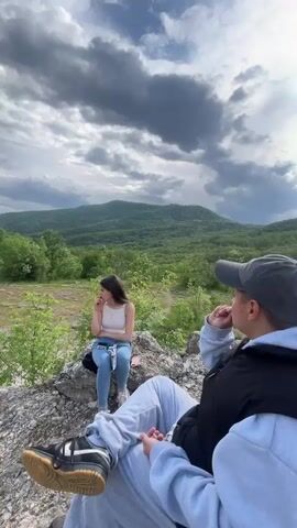 Two individuals are present in the video, a young woman and a young man, situated on a rocky outcrop. The woman is seated, wearing a white tank top and light blue jeans. She appears to be looking at her phone. The man, wearing a gray baseball cap and a dark jacket over a light gray hooded sweatshirt, is sitting beside her. His legs are extended, revealing black and white sneakers with tan soles. The camera angle is slightly above ground level, tilting upward. The background consists of a landscape with a verdant, forested mountain and a sky filled with a mix of clouds, including both dark storm clouds and patches of blue sky with scattered white clouds. The overall environment suggests a natural, outdoor setting.