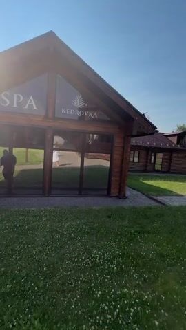 The video begins with a view of a wooden building with large windows, displaying the "KEDROVKA" logo. A person in a white robe is visible inside through the window. The camera pans to the right, revealing more of the wooden complex, a grassy area with a children's playground, and a clear blue sky. The camera then moves along a paved walkway, passing by another wooden structure with an outdoor seating area. A person in a white robe walks away from the camera down the path. The video continues to follow the paved path, which curves around a lush green hillside dotted with trees. The scene appears to be a resort or a similar recreational facility in a natural, wooded environment.