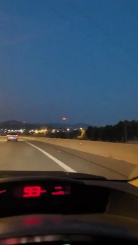 The video is taken from the perspective of a car driving on a highway at dusk. The dashboard displays the speed as 93. The road has white lane markings and orange reflectors on the side barrier. In the distance, there are streetlights and buildings, and a large orange moon is visible in the dark blue sky.