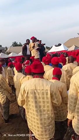 A large crowd of men are gathered outdoors, likely at an event or ceremony. Most of the men are wearing identical beige shirts with a grid pattern and red caps. Some have additional red necklaces. They are standing closely together, facing a small group of men on a raised platform. One of the men on the platform is speaking into a microphone while holding a paper. Other men on the platform are holding phones, possibly recording the event. In the background, there are white tents and a building. The sky is slightly overcast. The camera appears to be handheld and is moving slightly through the crowd. A watermark indicates the video was shot on a POWER P55.