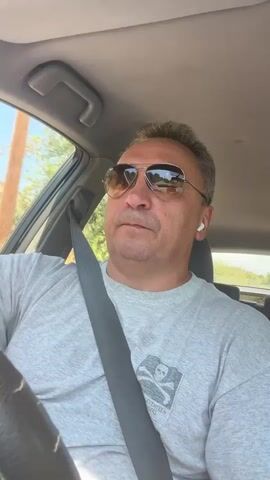 The video shows a man with short, grey hair and sunglasses wearing a grey t-shirt with a skull and crossbones graphic on it, driving a car. He has a seatbelt fastened across his chest and appears to be wearing white wireless earbuds. The interior of the car is visible, including the dashboard and a portion of the steering wheel. Outside the car, trees and foliage can be seen, suggesting a rural or suburban setting with a sunny sky. The camera is positioned inside the car, looking towards the driver. The man's gaze is directed forward, suggesting he is focused on the road.