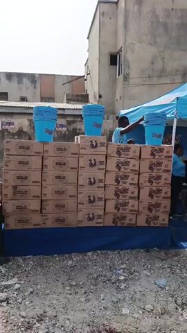 The video shows a small blue pop-up stall with a blue canopy. The canopy is branded with the words "GOLDEN MORN" in large white letters. The stall is constructed of wood. There are stacks of cardboard boxes with "GOLDEN MORN" printed on them. There are also stacks of blue buckets on top of the cardboard boxes. Several people are standing behind the stall. One person is a woman wearing a white hijab. A man in a red vest and black pants approaches the stall. Other people are visible in the background, milling around. The stall appears to be set up in a somewhat unpaved, dusty area near some buildings. There's a sign on the side of the stall that starts with the word "Disclaimer."