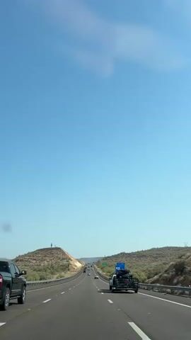 The video shows a view from a vehicle driving on a highway. The sky is clear and blue with some wispy clouds. On the left side of the highway, there is a hill with some sparse vegetation and two people standing on top. On the right side of the highway, there is more desert vegetation. There are several vehicles on the road, including the vehicle the video is being recorded from. One vehicle in front is a black pickup truck. Further ahead, another vehicle is a white pickup truck towing a trailer with something on it, possibly an ATV or dirt bike. There are also other cars and trucks in the distance. A blue highway sign with white lettering is visible on the right side of the road. The scene suggests a sunny day in a desert or arid environment.