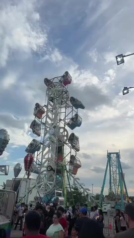 The video shows a crowded carnival scene. In the foreground, a tall, white amusement ride with multiple gondolas is visible. To the right of this ride, a teal-colored pendulum ride is also operating. People are milling around the base of the rides, and the sky is cloudy. There are lights on the rides, and the overall atmosphere appears lively. The camera pans slightly, showing more of the carnival attractions and the sky.