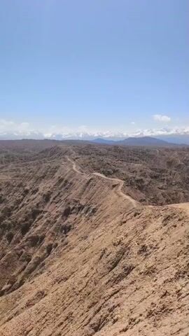 The video presents a panoramic view of a vast, arid, and rugged mountainous landscape. The foreground and mid-ground consist of numerous dry, eroded hills and ridges, predominantly in shades of brown, tan, and reddish-brown, indicative of a canyon or badlands-like environment. A narrow dirt path or track winds along one of these prominent ridges.

In the distant background, a majestic range of snow-capped mountains stretches across the horizon under a clear, bright blue sky with a few wispy clouds. As the video progresses, the camera slowly zooms in, revealing one or two small, indistinct figures walking or standing on the distant dirt path along the ridge, highlighting the immense scale of the natural environment. The lighting suggests it is daytime, likely mid-day, with strong, natural sunlight. The scene conveys a sense of wild, untouched nature and vastness, characteristic of regions like Ak Say, Kyrgyzstan. No visible text is present in the video.
