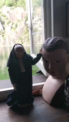 A static shot displays a tableau set on a wooden surface in front of a window. On the left, a doll depicting an elderly woman in a black nun's habit and glasses is positioned in a kneeling posture, her right hand resting on the head of a beige mannequin bust of a young boy with dark hair. To the right of the mannequin head, a dark bottle labeled "OLD PORT" is partially visible. A small, reddish object resembling a disembodied limb lies on the wooden surface near the nun doll. Through the window, a body of water, possibly a lake or quarry, is visible with light-colored rocky cliffs and green foliage on the far side.