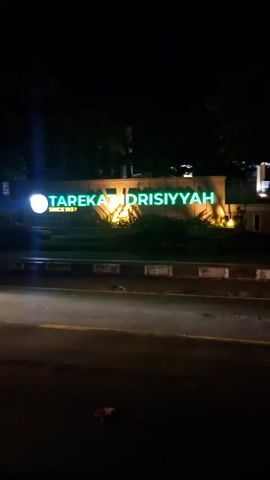 This video shows a nighttime street view of a building with illuminated green signage that reads "TAPEKA IDRISIYYAH". A car with its headlights on is moving past in the foreground, creating a blurred motion effect. In the background, a mosque is visible with illuminated minarets and a dome, as well as more green signage. There are also trees and streetlights in the scene.