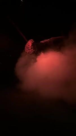 A globular object, possibly a light fixture or art installation, hangs suspended in darkness. It is entangled with what appears to be red twine or rope, creating a rough, textured surface. A thick plume of red smoke billows around the object, obscuring the background. The smoke is illuminated by the red light, casting a glow on the surroundings. In the distance, faint green lights flicker, suggesting a forest or outdoor setting. The scene is dark and atmospheric, with the red light and smoke creating a dramatic effect.