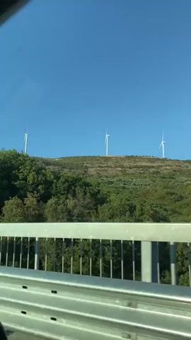 A video taken from a moving vehicle, looking out over a metal railing and guardrail. The scene is a hilly landscape with lush green trees and vegetation. In the distance, on the crest of a hill, are several wind turbines silhouetted against a clear blue sky. The video captures the movement of the vehicle, causing the scenery to pan from left to right.