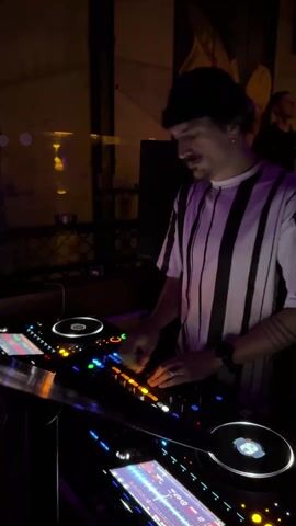 In a dimly lit bar, a man in a black beanie and a white t-shirt with black vertical stripes is DJing. He has a mustache and is wearing rings on his fingers and a bracelet on his left wrist. He stands behind a DJ console with two turntables and a mixer, using his hands to adjust the controls. The console is lit up with blue and orange lights. In the background, there's a dark wall with the name of the bar written in white letters and a dark-haired man is looking up. The light in the bar is changing colors from yellow to pink to purple.