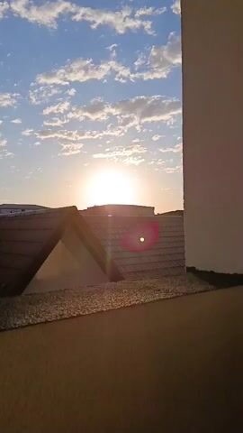 A video shows a sunset over a residential area. The sun is visible in the sky, partially obscured by clouds. In the foreground, there are rooftops of houses, with one house featuring a triangular gable. The sky is blue with scattered white clouds. The sun casts a warm glow over the scene, and there is a lens flare visible from the sun. The video pans slightly to the right, revealing more houses and a power line in the distance.