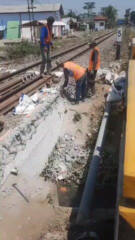 Three construction workers, wearing orange safety vests and blue shirts, are working on railroad tracks. One worker on the left stands with their back to the camera. The middle worker is kneeling and appears to be using a jackhammer on a concrete structure. The third worker stands behind the kneeling worker, observing the process. The environment shows dislodged concrete, rubble, building materials in white bags, and railroad tracks extending into the distance. The scene is outdoors under a bright, sunny sky.