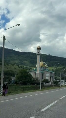 The video shows a mosque with a golden dome and a tall minaret, situated in a valley with lush green hillsides under a cloudy sky. A paved road runs in the foreground, and power lines are visible overhead. The camera appears to be mounted on a moving vehicle, likely a car, as indicated by the perspective and slight motion blur. The mosque is a modern building with light-colored walls and aqua-colored accents on the roof. The minaret is tall and slender, also topped with a golden dome. Trees and some smaller buildings are visible around the mosque. The overall scene depicts a serene and picturesque landscape with religious architecture.