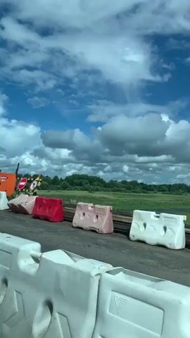 The video pans across a construction site. In the foreground, there are white and red plastic barriers. Behind them, on the left, is an orange generator. Further back, there are more plastic barriers and rubble. A blue fence with wooden posts runs across the middle of the frame, separating the construction area from a field and a body of water. The sky is blue with scattered white clouds.