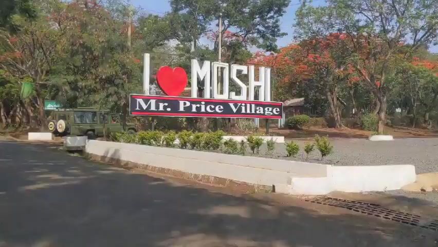 The video begins with a static shot of a sign that reads "I [Heart Symbol] MOSHI" above a sign that reads "Mr. Price Village." The camera then pans to the right showing an olive green jeep parked near a smaller sign. The camera then moves to the perspective of the inside of a moving vehicle, presumably a tuk-tuk, driving down a paved road lined with trees. Another white vehicle is seen driving in the distance. A black tuk-tuk drives by in the opposite direction. The view then shifts to the back window of the red tuk-tuk.