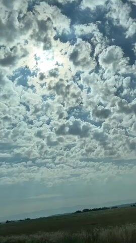 This is a vertical video captured from a moving vehicle, likely a car given the lower-left corner framing. The scene is dominated by a vast sky filled with a dense, textured layer of cumulus clouds. Sunlight filters through, creating bright patches and casting shadows, suggesting a partly cloudy day. The sun itself is partially obscured by the clouds, indicating it is either mid-morning or mid-afternoon.

Below the dramatic sky lies a wide expanse of green fields, appearing to be agricultural land with rows of crops. The fields stretch towards a distant horizon where rolling hills or mountains are faintly visible. A thin line of trees marks the boundary of the fields in places, and some indistinct structures can be seen along the horizon.

The overall mood is serene and expansive, with the vastness of the sky and the open fields evoking a sense of peace and natural beauty. There is no visible text in the video. The provided location context of Sântionlunca, Romania, places this rural landscape within that region. The activity is simply the observation of the natural scenery as the viewer (or camera) moves through the landscape.
