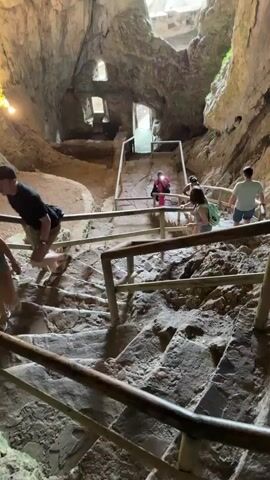 The video begins inside a large cave with people walking down a staircase. The cave has rough, textured rock walls and a large opening at the top that lets in natural light. As the camera moves forward and closer to the opening, the view shifts to a landscape of a village nestled in a valley surrounded by lush green trees and hills. The sun is shining brightly, illuminating the scene. The video concludes with a clear view of the village and its surroundings from the cave opening.