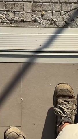 This media file depicts an outdoor scene under a clear blue sky, likely in the morning or late afternoon due to the angle of the sun and the shadows. The foreground shows a tiled patio area that appears to be under construction or renovation. Some of the tiles are broken, and there is debris on the ground, suggesting recent work. A white trim piece runs horizontally across the image, possibly a threshold or the base of a door frame. A person's foot, wearing a work boot, is visible at the bottom right corner, indicating someone is present and possibly involved in the construction.

Beyond the patio, a scaffolding structure is set up, with stacks of terracotta roof tiles on either side. These tiles are a common sight in Mediterranean regions and further hint at renovation or building work. In the background, lush green vegetation and several tall palm trees are visible, characteristic of a warm, sunny climate like Italy. The sun is shining brightly, casting distinct shadows from the scaffolding and palm trees onto the tiled ground. The overall impression is one of an outdoor construction site bathed in pleasant sunlight, with a clear view of the surrounding landscape. There are no visible text elements in the media.