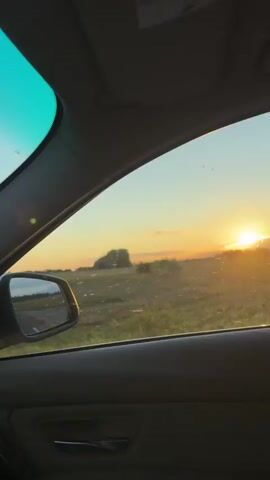 The video captures a view from inside a car looking out the passenger window at a sunset. The sun is visible on the horizon, casting a warm glow over a grassy field dotted with trees. A side-view mirror is visible on the left, reflecting the road ahead. The interior of the car is dimly lit, with parts of the dashboard and door panel visible. The camera pans slightly, revealing more of the trees and the sky as the sun continues to set. The overall scene conveys a sense of a peaceful drive through the countryside.