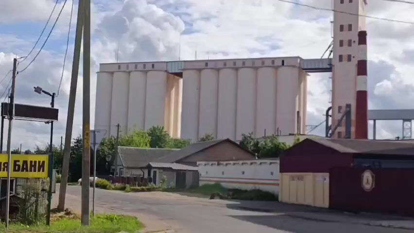 The video shows a long shot of an industrial facility on a cloudy day. The facility appears to be a grain elevator or storage facility, with several large, cylindrical silos. A tall, narrow tower with red and white stripes rises from the center of the facility, likely a smokestack or processing tower. Several connecting structures and conveyor belts are visible between the silos and the tower. In the foreground, there are some smaller buildings, a fence, and a street. Power lines cross the frame. The camera pans slightly to the right throughout the clip.